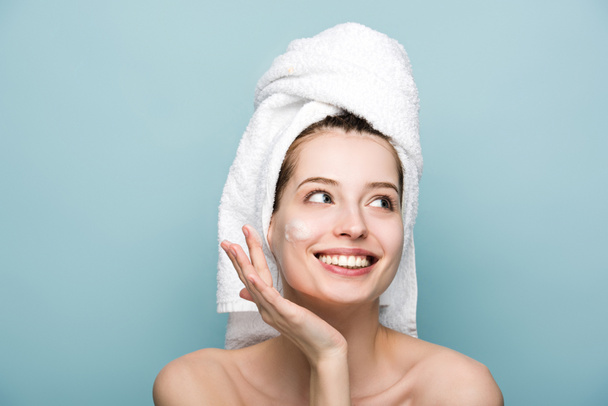happy attractive girl applying moisturizing facial mask while looking away isolated on blue - Zdjęcie, obraz