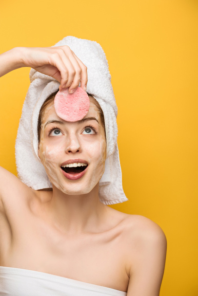 vzrušená dívka s hydratační obličejová maska při pohledu na kosmetické houbě izolované na žluté - Fotografie, Obrázek