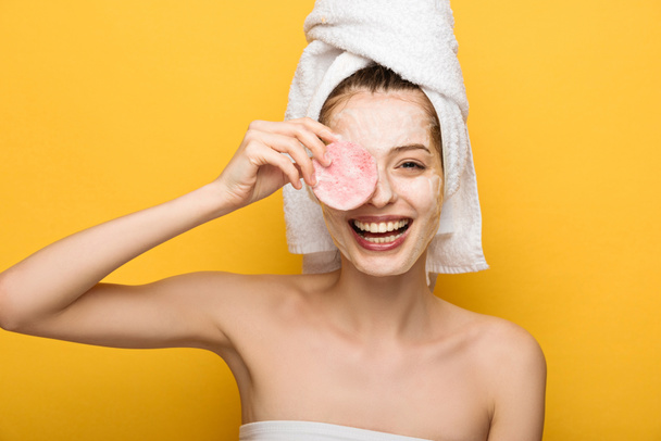 fröhliches Mädchen mit feuchtigkeitsspendender Gesichtsmaske, die Augen mit Kosmetikschwamm auf gelbem Hintergrund bedeckt - Foto, Bild