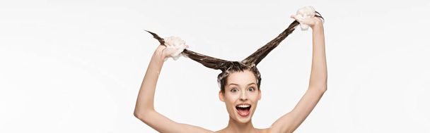 Panoramaaufnahme eines aufgeregten Mädchens, das Spaß hat, während es lange Haare isoliert auf Weiß wäscht  - Foto, Bild