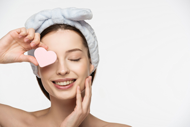 happy girl covering eye with heart-shaped cosmetic sponge while touching face isolated on white - Φωτογραφία, εικόνα
