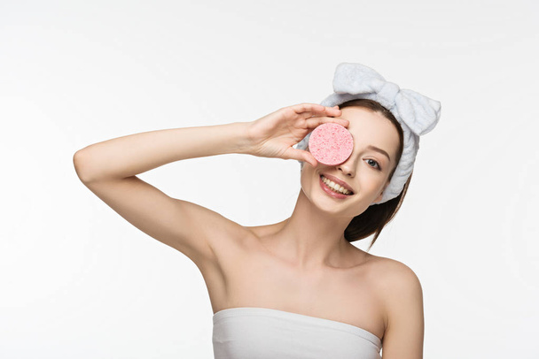 happy girl covering eye with cosmetic sponge while looking at camera isolated on white - Foto, afbeelding