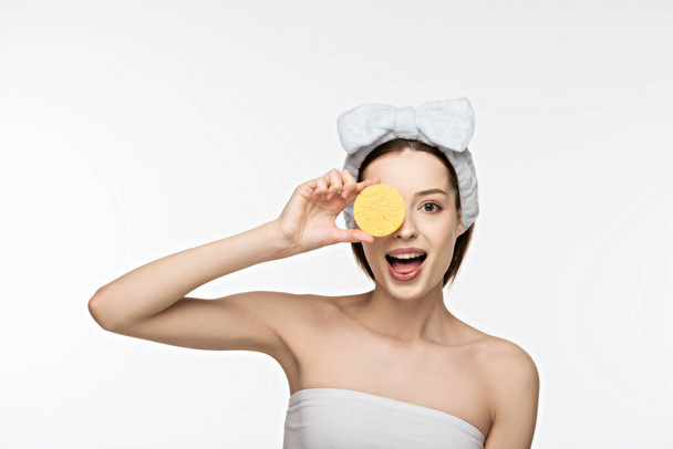 cheerful girl covering eye with cosmetic sponge while looking at camera isolated on white - Photo, Image