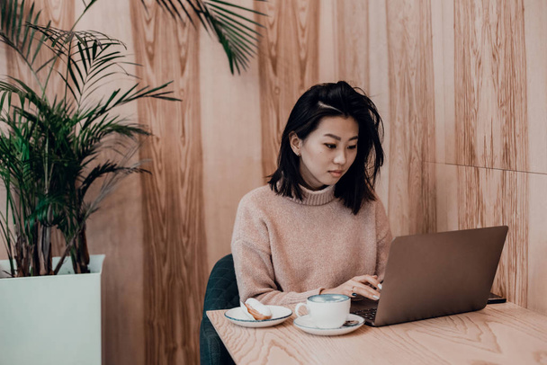 Милая взрослая девушка играет в ноутбуке в кофейне. Азиатка, работающая в кафе за ноутбуком. Концепция фриланса - Фото, изображение