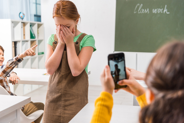 selective focus of schoolboy pointing with finger at upset kid while schoolgirl taking photo, cyberbullying concept  - Photo, Image