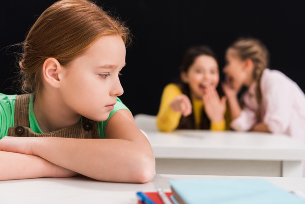selective focus of upset schoolkid sitting near classmates laughing and gossiping in classroom isolated on black, bullying concept  - Photo, Image