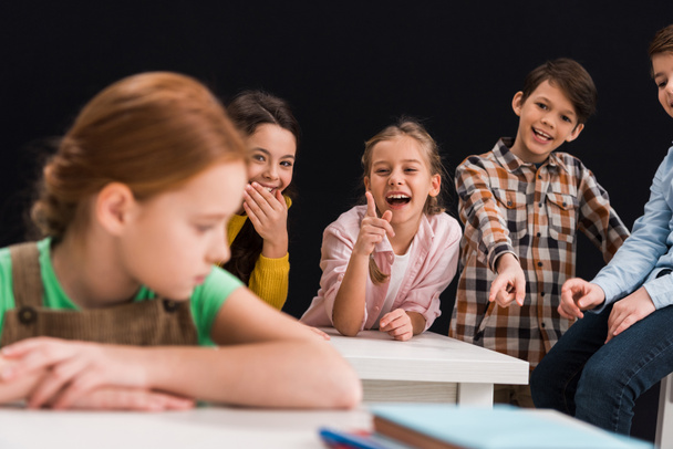 selective focus of classmates laughing and pointing with fingers at schoolgirl isolated on black, bullying concept  - Photo, Image