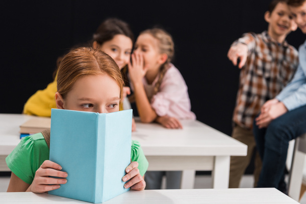 selective focus of upset schoolkid covering face with book near classmates gossiping and pointing with finger isolated on black, bullying concept  - Photo, Image