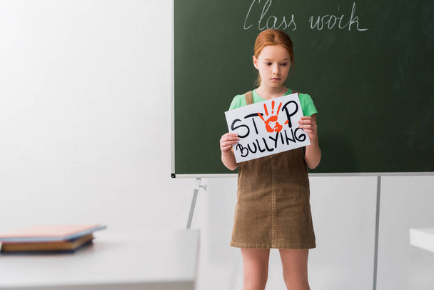 selective focus of sad kid holding placard with stop bullying lettering in classroom  - Valokuva, kuva