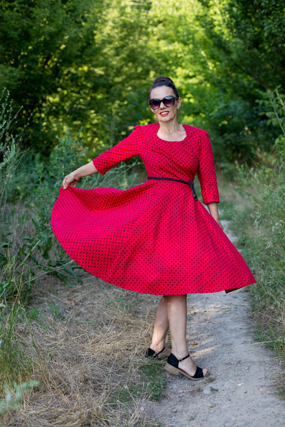 Mulher feliz meia idade girar em beautfiul vermelho retro vintage midi vestido com pontos e cinto, vestindo óculos e sorrir ao ar livre, elegância feminina e beleza
 - Foto, Imagem