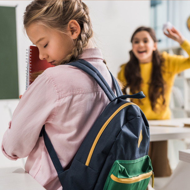selective focus of bullied schoolgirl with backpack holding notebooks near cruel schoolkid - Фото, изображение