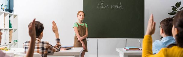 colpo panoramico di scolaretta in piedi vicino alla lavagna con scritte di lavoro di classe e compagni di classe con le mani alzate
  - Foto, immagini