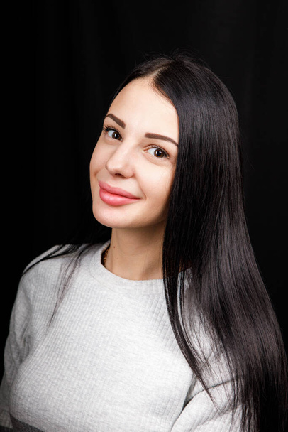 Portrait of serious beautiful female with black hair, has minimal makeup, looks calmly at camera, wears white jumper, stands against black background, being deep in thoughts - Fotoğraf, Görsel