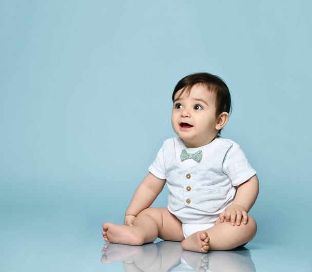 Little toddler in white bodysuit as a vest with bow-tie, barefoot. He is sitting on the floor against blue background. Close up - Foto, imagen