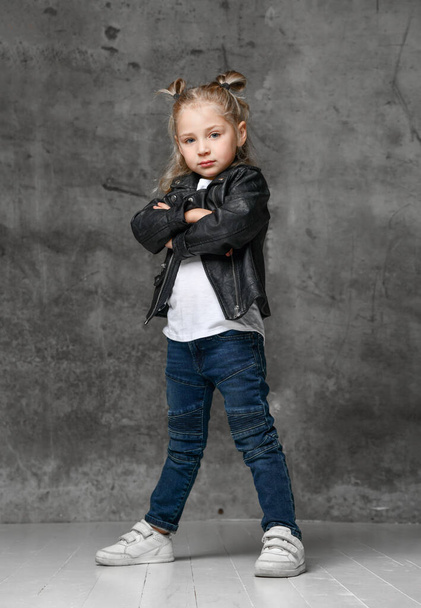 Little cute smiling blond girl in stylish rock style black leather jacket, jeans and white sneakers standing in confident pose - Fotó, kép