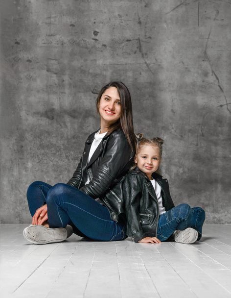 Positive young woman and cute little girl in similar rock style clothing and black leather jackets posing over grey background - Foto, immagini