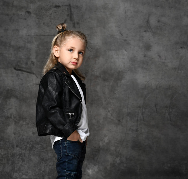 Little cute smiling blond girl in stylish rock style black leather jacket and blue jeans standing over grey concrete background - Fotografie, Obrázek