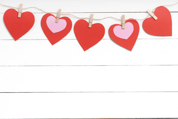 Μανταλάκια με κόκκινες καρδιές κρεμασμένες σε σχοινί πίσω από λευκή ξύλινη επιφάνεια. - Φωτογραφία, εικόνα