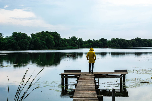 Стильный молодой человек в желтом водонепроницаемом плаще стоит на деревянном пирсе у озера или реки
 - Фото, изображение