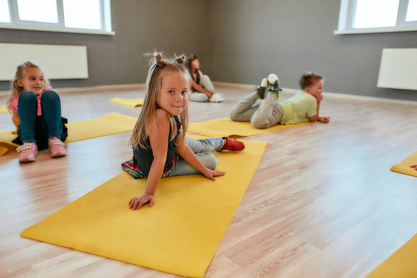 Portrait of a cute little girl sitting on yellow yoga mat and looking at camera with smile. Group of children lying on yoga mats and relaxing after gymnastic class - Fotografie, Obrázek