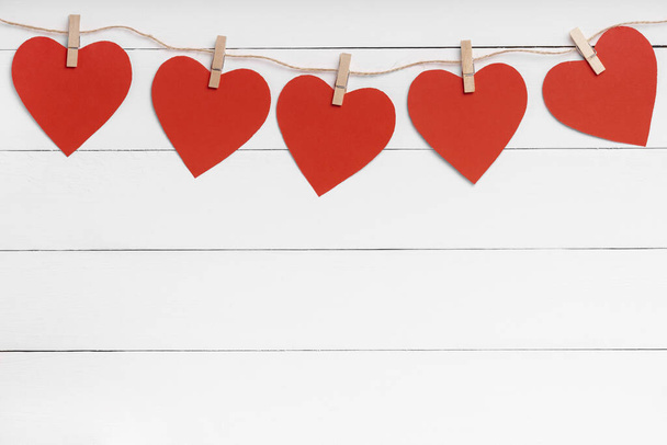 Μανταλάκια με κόκκινες καρδιές κρεμασμένες σε σχοινί πίσω από λευκή ξύλινη επιφάνεια. - Φωτογραφία, εικόνα
