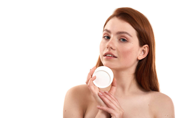 Taking care of her sensitive skin. Young attractive red-haired girl with freckles demonstrating cosmetic product and looking at camera while standing in studio against white background - Fotó, kép