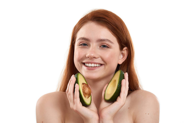 Perfect for my skin. Portrait of young and happy red-haired girl with freckles on face holding two pieces of avocado and smiling at camera while standing against white background - Fotó, kép