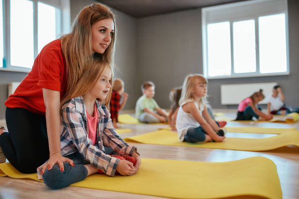 Stretching Practice. Side view of a cute little girl doing butterfly exercise with female trainer. Group of children sitting on the floor and stretching muscles. Dance studio - Foto, imagen