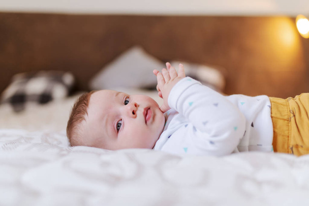 Widok z boku słodkie kaukaskie sześć miesięcy chłopiec leżący na łóżku w sypialni i zabawy rękami podczas patrząc na aparat. - Zdjęcie, obraz
