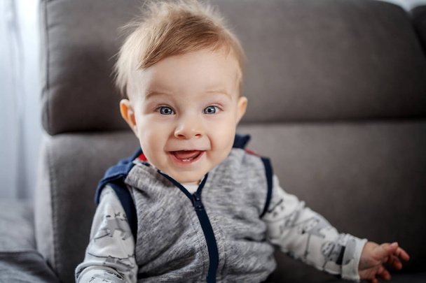 Vrolijke blanke kleine jongen met grote blauwe ogen zittend op de bank in de woonkamer, glimlachend en kijkend naar de camera - Foto, afbeelding