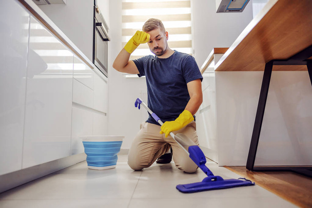 Joven hombre barbudo digno de orden que se arrodilla, limpiando el sudor y tratando de limpiar el piso sucio en la cocina
. - Foto, imagen