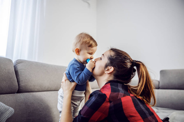 Young attractive brunette with ponytail dressed casual holding her only son and playing with him. Toddler chewing teether and looking at mom while standing on sofa. - Fotó, kép