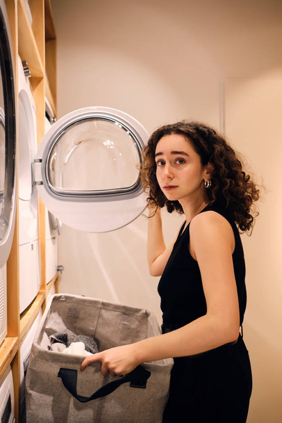 Gyönyörű lány figyelmes nézi a kamera betöltése ruhák mosógép a modern önkiszolgáló mosoda - Fotó, kép