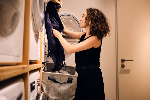 Гарненька брюнетка дівчина щасливо завантажує одяг в пральну машину в пральні самообслуговування
 - Фото, зображення