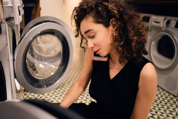 Όμορφη μελαχρινή κοπέλα μιλάμε για smartphone πλύσιμο ρούχων στη σύγχρονη αυτο-υπηρεσία πλυντηρίου - Φωτογραφία, εικόνα