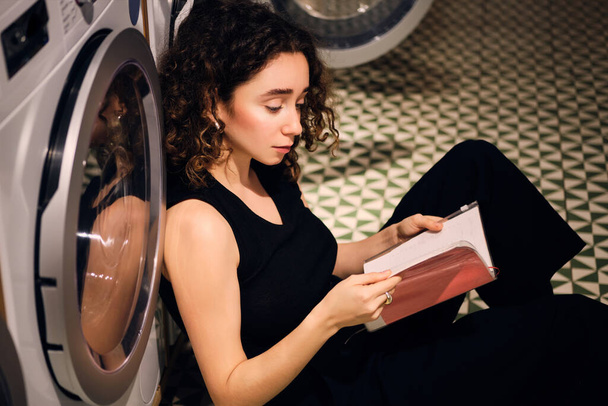 Вид сбоку привлекательной девушки, мечтающей читать книгу в ожидании стирки в современной лавке самообслуживания
 - Фото, изображение