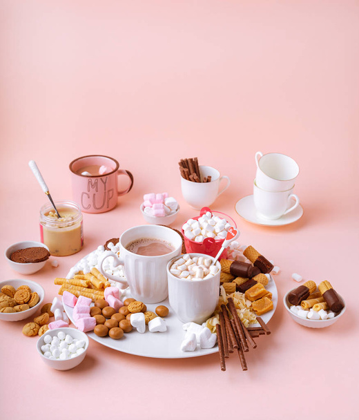 Gorący napój czekoladowy w kubkach, słodycze asortyment pianek, czekoladki i ciasteczka na różowym tle - Zdjęcie, obraz
