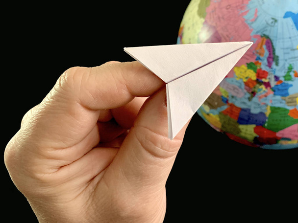 Papírová rovina v ruce na pozadí koule, černé pozadí. Let letadla po celém světě. Muž ukazuje rukou na glóbus. Koncept: cestování, cestovní ruch, - Fotografie, Obrázek