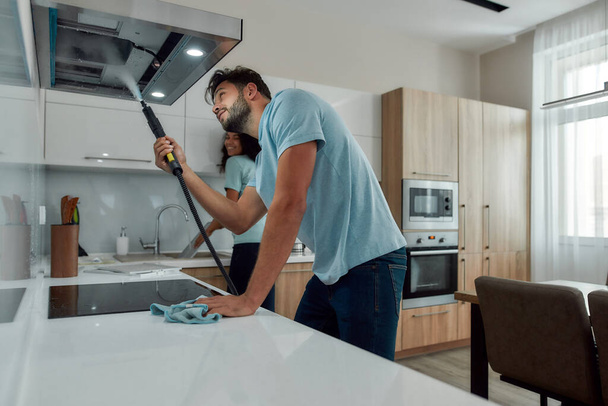 Young caucasian man in uniform cleaning kitchen range hood with steam cleaner while his smiling female colleague washing something in the sink on the background. Cleaning services concept - Fotoğraf, Görsel