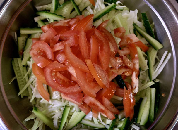 Πράσινη σαλάτα λάχανο, αγγούρια και ντομάτες, σε μαύρο φόντο. Κοπή λαχανικών σε ένα μεταλλικό μπολ. Ψιλοκομμένα λαχανικά για σαλάτα. Έννοια: φυτική διατροφή, υγιεινή διατροφή. - Φωτογραφία, εικόνα