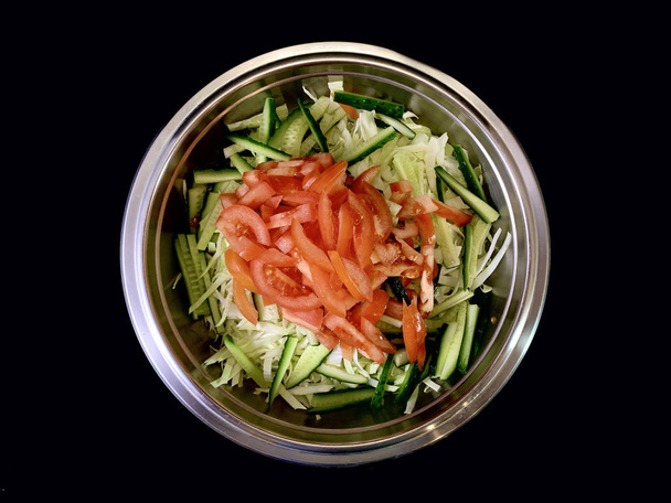 Green salad of cabbage, cucumbers and tomatoes, on a black background. Cutting vegetables in a metal bowl. Finely chopped vegetables for salad. Concept: vegetable diet, healthy nutrition. - Valokuva, kuva