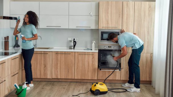 Keeping your house clean. Two young professional cleaners in uniform working together in the kitchen. Young caucasian man using steam cleaner, happy afro american woman cleaning kitchen range hood. - 写真・画像