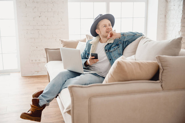 Hombre barbudo compañero de trabajo hipster en sombrero de trabajo sala de estar casa. Hombre sentado en el sofá utilizando el ordenador portátil y el teléfono móvil. Fondo de luz borrosa
 - Foto, imagen