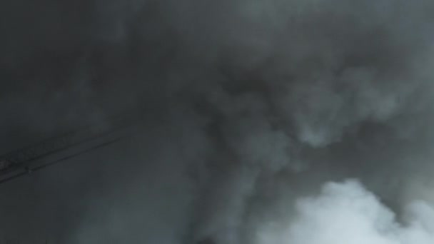 Egy azonosítatlan tűzoltó jelenik meg a füstből, ahol hatalmas tűzzel küzd a tűzoltóautó létrájának végéről, és lentről lövi a vizet.. - Felvétel, videó