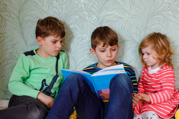 Two school kids boys and little toddler girl reading together book. Older brother holding book and read aloud for his sibling. Family spending time together. Three children. - Photo, Image