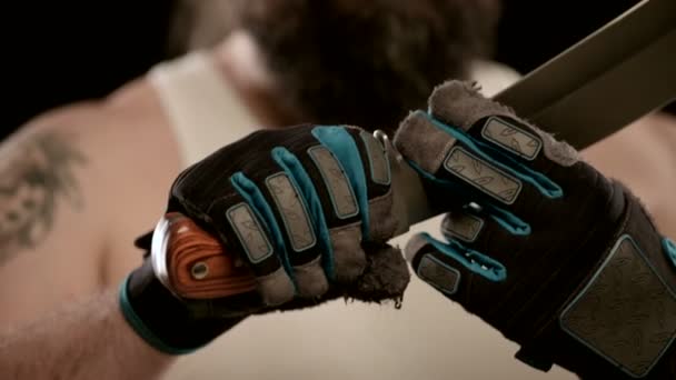 Man with beard, gloves in black studio, runs his fingers along blade of machete. - Πλάνα, βίντεο