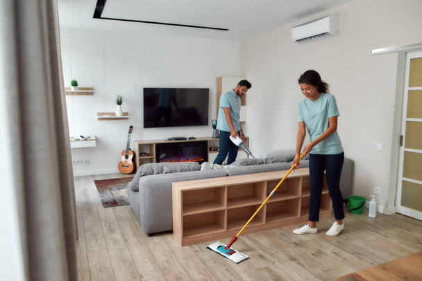 Cleaning team. Two professional cleaners in uniform working together in the living room. Young afro american woman cleaning floor with mop while her male coworker cleaning sofa with handheld vacuum - 写真・画像