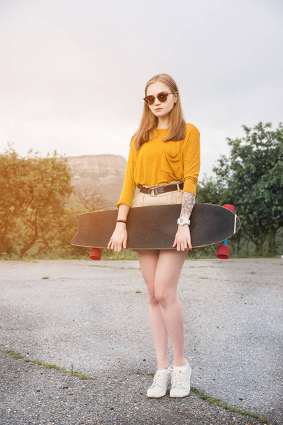 Beautiful and fashionable young woman in sunglasses and with a tattoo poses with a skateboard or longboard against the sunset sky - Photo, Image