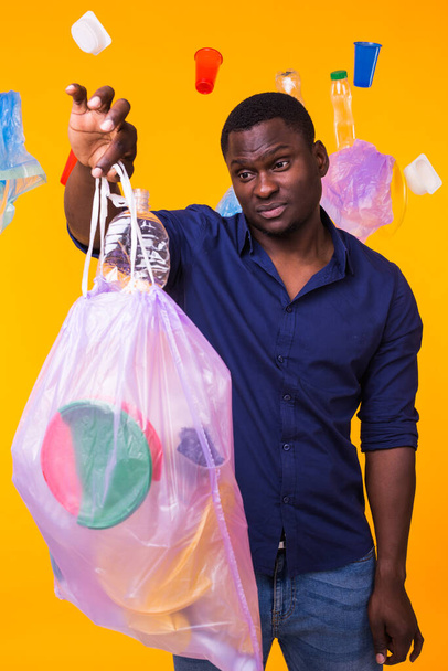ゴミの問題、プラスチックのリサイクル、汚染と環境の概念-黄色の背景にゴミ袋を運ぶ混乱した男 - 写真・画像