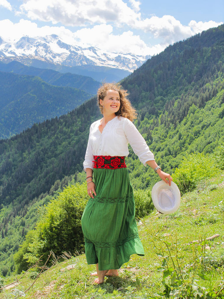 Elegancka kobieta w zielonej spódnicy w górach. Boso na trawie. Szczęśliwa dziewczyna z falowanymi włosami z kapeluszem. - Zdjęcie, obraz